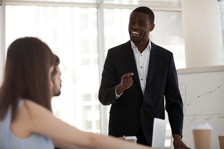 Diverse millennial people at meeting in office boardroom gathered together negotiate solve business matters. Black positive experienced coach speaking discussing with company employees during lectureの写真素材