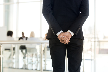 View from distance diverse coworkers company staff negotiating behind a closed glass door, focus on businessman wearing suit standing with crumpled paper in folded hands behind his back, rear viewの写真素材