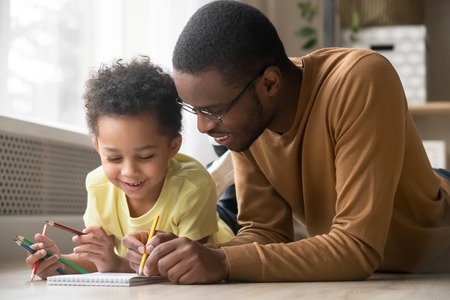 African dad and little creative toddler kid son draw with colored pencils lying on warm floor together, black father baby sitter teaching help child boy learning playing at home, babysitting conceptの写真素材
