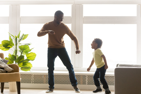 Happy african american dad and little toddler son dancing in living room together, funny kid boy having fun with black father laughing moving to music, daddy baby sitter child play together at homeの写真素材