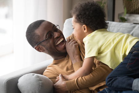 Happy african american dad laughing playing with little toddler son together, playful mixed race kid boy tickling black father cuddling enjoying moment of love having fun with daddy at home on couchの写真素材