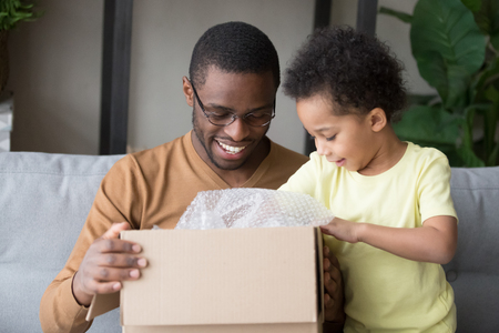 Happy black father and little kid son opening cardboard box looking inside, african american dad with cute child toddler clients receiving carton package, post mail parcel delivery service conceptの写真素材