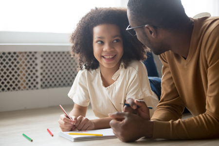 Happy cute mixed race kid daughter looking at dad play draw with colored pencils lying on warm floor together, african father baby sitter teaching child girl having fun at home, creative family hobbyの写真素材