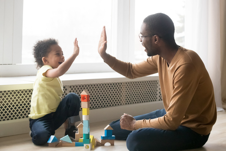 Happy black dad and cute toddler son giving high five playing with wooden blocks on floor at home, little kid boy enjoying game with african father, daddy child clapping hands having fun togetherの写真素材