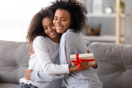 Relative people sitting on couch at home, african mother embracing teen daughter congratulating lovely child with birthday or special occasion. Love and attention, best wishes and life events conceptの写真素材