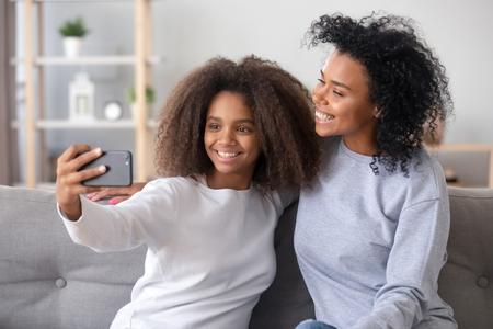 African older younger sisters sitting on couch spending time at home hold smartphone making video call talking with friends. Mother and teen daughter taking selfie photo using mobile phone applicationの写真素材