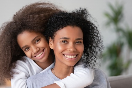Close up african mother piggyback daughter smiling looking at camera feels happy. Younger embracing from behind older sister. New mommy for adopted kid, good relations between parent and child conceptの写真素材