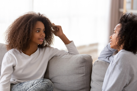African adolescent girl sitting together with older sister friend or friendly nanny chatting sharing secrets. Young mother listening teenage child spending free time at home. Trust and support conceptの写真素材