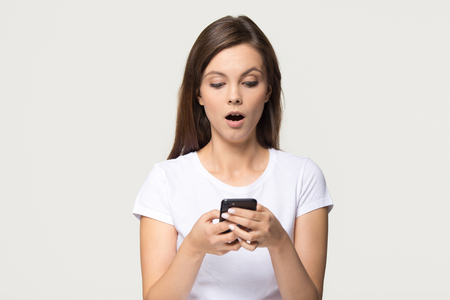 Shocked teen girl holding phone feeling amazed about unexpected mobile message social media news, surprised stunned young woman looking at smartphone screen isolated on white grey studio backgroundの写真素材
