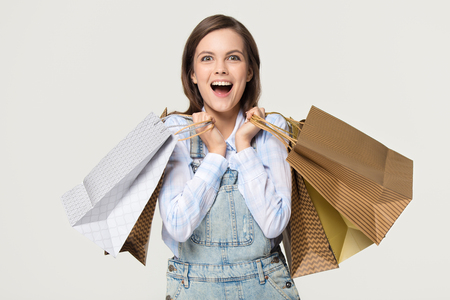 Excited happy teen girl shopper holding many paper bags enjoy fashion sale, overjoyed ecstatic young woman model customer shopaholic carrying shop purchases isolated on white grey studio backgroundの写真素材