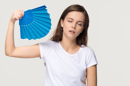 Tired young woman feel overheated suffering from heat stroke high temperature sweating problem, sweaty girl holding waving fan cooling in hot summer weather isolated on white grey studio backgroundの写真素材