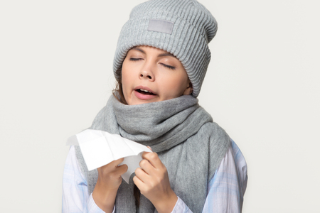 Sick young woman in winter scarf and hat holding handkerchief sneezing got flu cold symptoms, ill teen girl coughing isolated on white grey studio background, flue grippe rhinitis allergy conceptの写真素材