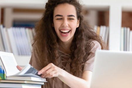 Happy teen girl college student laughing study with books and laptop in library, cheerful teenager having fun enjoy reading funny online content social media computer news or watching computer videoの写真素材