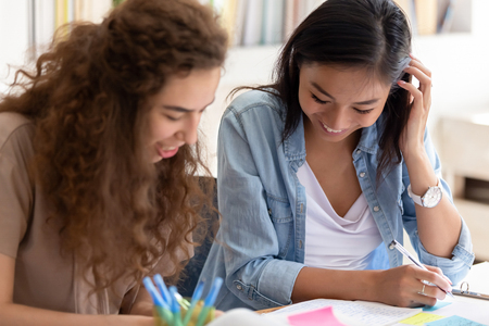 Two happy diverse girls college friends study together in campus, teen asian and caucasian female students team talking laughing helping in preparing university creative research project make notesの写真素材