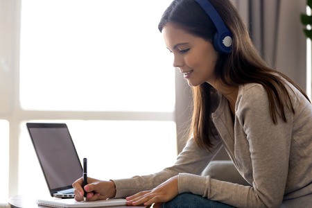 Focused woman in headphones writing notes, using laptop, learning language or preparing to exam, watching webinar, listening lecture, online education concept, interpreter working at homeの写真素材