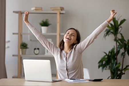 Excited smiling woman with closed eyes stretching hands at workplace after finish work, relaxing, doing easy exercise during break, happy girl enjoying free time, celebrating successの写真素材