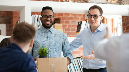 Black man having first working day getting acquainted with colleagues standing in office in front of workmates. Boss introducing employee, newcomer holds box with belongings starting career in company