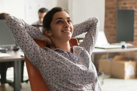 Dreamy happy indian businesswoman relaxing taking break at work, calm smiling hindu lady resting sitting in ergonomic chair dreaming hands behind head breathing fresh air feel no stress enjoy balanceの写真素材