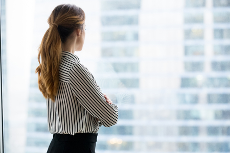Rear back view at confident rich businesswoman looking forward through glass window of skyscraper building dreaming of success thinking of future business vision and new goals enjoying city view.の写真素材