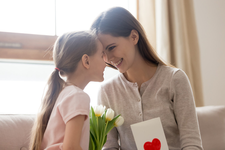 Happy mom and little kid daughter bonding celebrating mothers day, cute child girl making surprise congratulating mom with birthday holding spring flowers and greeting card, family love celebrationの写真素材