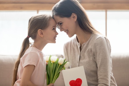 Cute little kid daughter making surprise congratulating happy mom with mothers day birthday 8 march presenting flowers greeting card, smiling loving affectionate family mum and child girl, side viewの写真素材