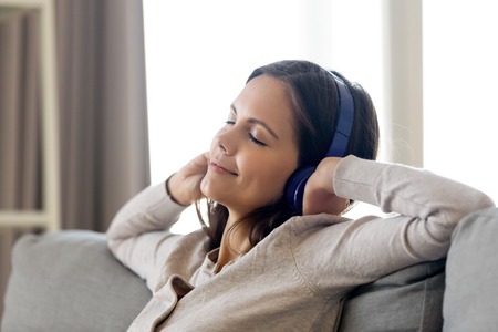 Smiling young woman leaning on sofa wearing headphones closed her eyes listening music enjoy favourite song audio track feels good. Leisure activities, hobby and free time relaxation at home conceptの写真素材