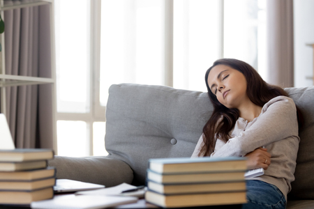 Pile heap of books educational material on table, focus on tired student sleeping leaning on sofa in living room at home fatigued of learning exam homework preparations, girl bored of studying conceptの写真素材