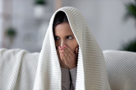 Woman sitting on couch covered wrapped with plaid warming up hands with breath feels frozen discomfort at cold home, seasonal weather period, unhealthy sick ill girl, central heating problems conceptの写真素材