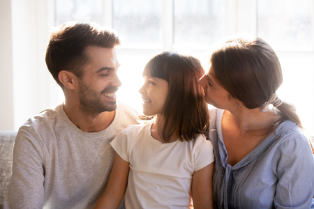 Attractive happy caucasian parents sitting on sofa with adorable small preschool daughter, family enjoy time with each other at home, adoption custody and upbringing care protection and love conceptの写真素材