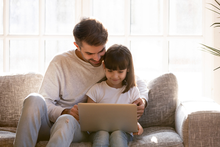 Father and little daughter sitting on sofa hold laptop having fun watching cartoons videos online, surfing internet, reading media, dad teach kid, modern wireless technology leisure activities conceptの写真素材