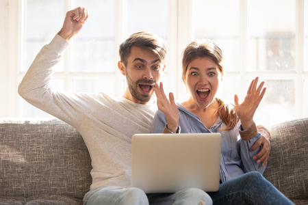Emotional couple sitting on couch using laptop looking at device screen making big eyes open mouth feeling triumph received great news online lottery win, cheap flights tickets, huge discounts conceptの写真素材