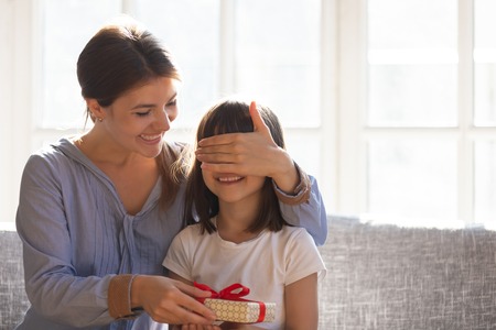 Smiling young loving mother cover daughter eyes with hand give her surprise, family celebrating child birthday holiday life event feels excited and happy, gift box as symbol of love care and attentionの写真素材