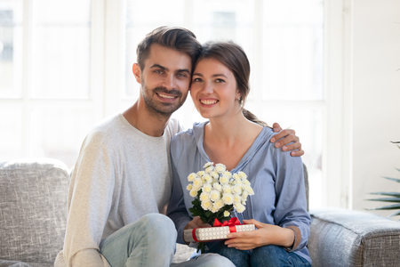 Happy couple sitting on sofa smile looking at camera, husband presented wife flowers gift box family celebrating eight 8 march International Womens Day birthday wedding anniversary life events conceptの写真素材
