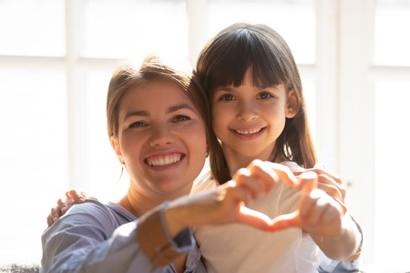 Head shot portrait mother and daughter make heart shape with hands fingers hugging sitting on couch sunlight through the window. Adoption and custody, health medical pediatric insurance, love conceptの写真素材