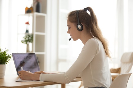 Student woman in headset with microphone writing notes, talking, using laptop at workplace, online distance learning language, call center operator working at home, e-learning concept side viewの写真素材