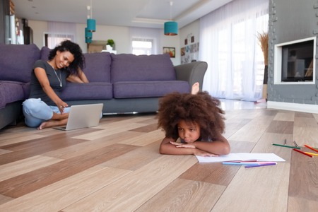 Upset African American girl feeling bored while mother using laptop, black woman chatting in social network, watching video online, ignoring frustrated daughter lying on warm floorの写真素材