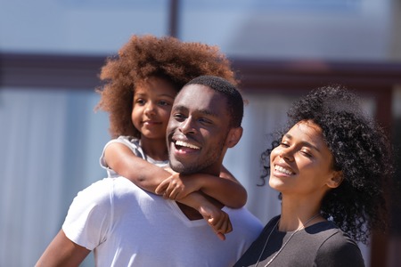 Happy African American family with daughter standing outdoor in yard of new house, smiling father holding little girl on back, piggyback, wife and husband with child having fun together, close upの写真素材
