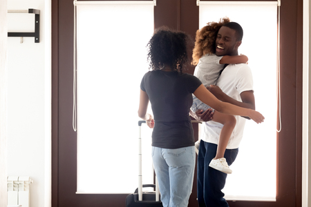 Happy African American father returning after long business trip, standing in entrance hall with luggage, holding little daughter in arms, excited wife welcoming back husband, huggingの写真素材