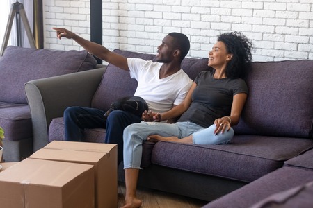 Happy African American couple planning renovation in new house, sitting on comfortable sofa with dog, excited man sharing ideas with attractive woman in living room, boxes with belongingsの写真素材