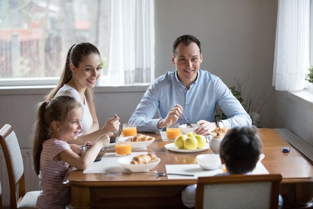 Happy family with children eating morning breakfast at kitchen. Family sitting at dining table at home. Mother and father smiling looking at funny son, daughter laughing brother refuse to eat mealの写真素材
