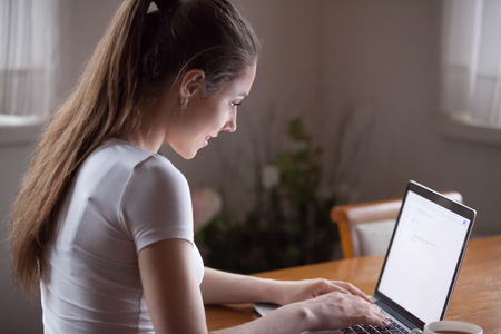 Smiling millennial woman typing email on laptop working at home. Female sitting at desk writing letter on computer looking at screen. Side view, profile look. Remote work, chatting online conceptの写真素材