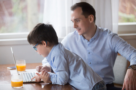 Father and son playing computer game on weekend at home. Family leisure time, gadget addiction concept. Smiling husband sitting work on laptop, kid in glasses look at monitor, poor eyesight problemの写真素材