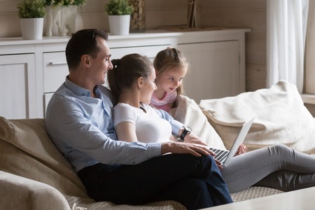 Family relaxing on sofa at home watching movie on computer. Happy people spending time together at home. Mother, father and daughter have leisure time on couch with laptop. Technology, gadgets conceptの写真素材