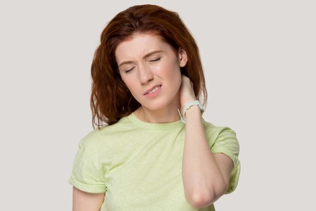 Exhausted red-haired young woman isolated on grey studio background massage neck having pain, tired redhead girl suffer from body spasm or strain, feel unwell from health problems. Bad posture conceptの写真素材