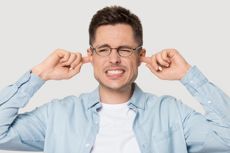 Annoyed Caucasian man wearing glasses isolated on grey studio background avoid hear bothering loud noise, irritated young male in spectacles plug fingers cover ears refuse listen to stressful soundの写真素材