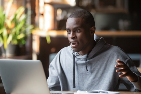 African millennial man sitting at table in cafe or office looking on computer screen feels amazed excited surprised by unexpected good news win result online unbelievable fantastic email offer concept.の写真素材