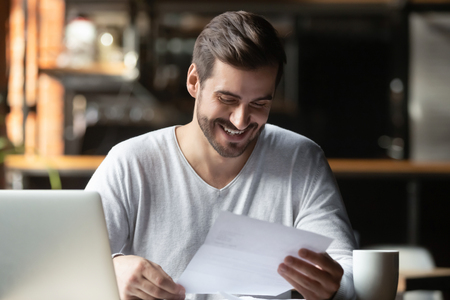 Satisfied millennial businessman sitting in cafe near computer holding paper reading letter feels happy overjoyed concept of getting notification bank loan approval, credit business, deal confirmationの写真素材