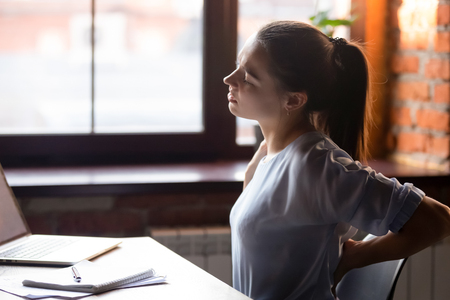 Side view young woman sitting at table working using computer take break touching massaging lower back feels discomfort after long sedentary studying, poor posture, incorrect stooped position concept