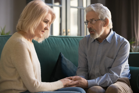 Grey haired husband comforting upset wife, expressing sympathy and understanding, sorry, mature couple holding hands, showing love and support, middle aged family sitting on comfortable sofa at homeの写真素材