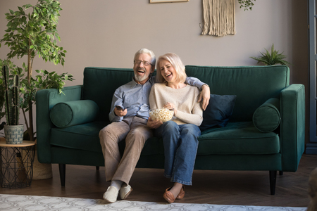 Happy aged couple, wife and husband watching tv, comedy movie and eating popcorn snack, laughing, sitting on cozy couch at home, mature family, man and woman enjoying free time, weekend togetherの写真素材